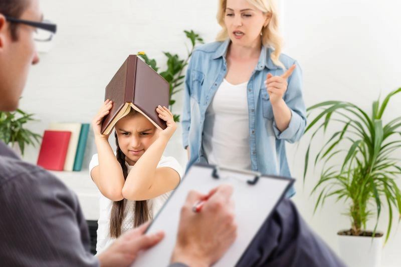 A child sitting with her hands on her head, appearing distressed while an adult woman scolds her. A child behavioral therapist can assist in addressing emotional distress and teaching healthy coping strategies for both children and parents.