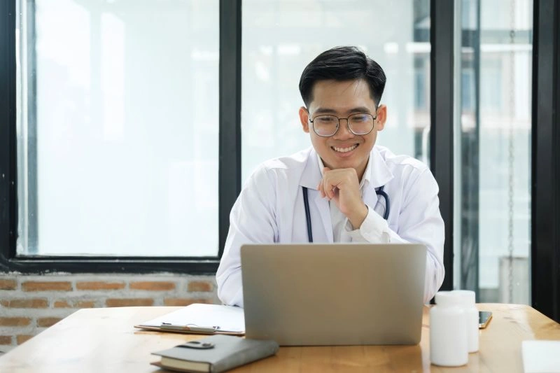 A smiling male psychiatrist in a white coat working on a laptop, possibly conducting an online consultation. Online psychiatrists can prescribe medications through virtual appointments, providing accessible mental health care.