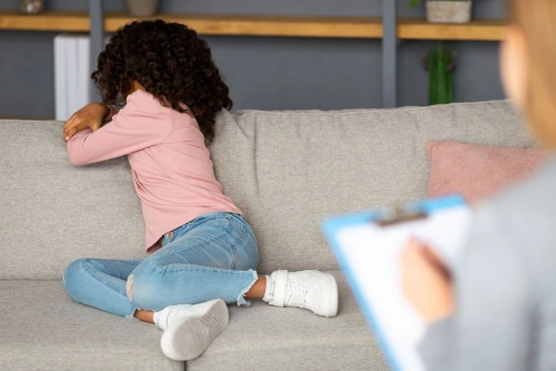 A young child sitting on a couch with their back turned and hands over their face, displaying signs of emotional distress during a session with a child psychiatrist. The therapist is taking notes, focusing on understanding the child's concerns.