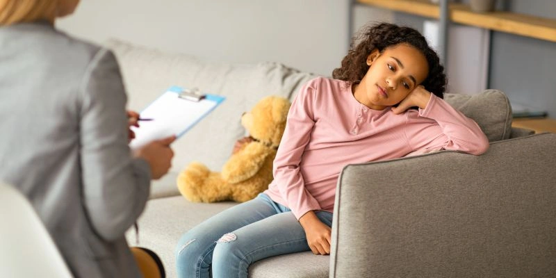 A child slumped on a couch, looking disinterested or disengaged while a therapist takes notes, reflecting the emotional challenges addressed in child psychiatry. The presence of a teddy bear suggests a child-friendly environment.