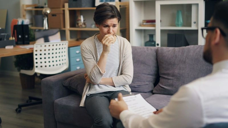 A woman sitting on a couch, anxiously listening to a therapist, showing the vulnerability often associated with trauma from childhood. The image depicts a common scenario in therapeutic settings addressing childhood trauma.